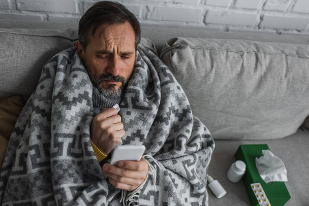 Mann mit warmer Decke bedeckt und schaut auf Smartphone, während er in der Nähe von Behältern mit Behandlung und Papierservietten sitzt - Foto, Bild