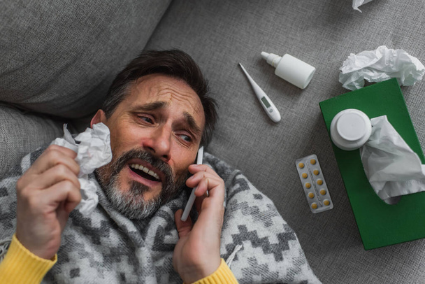 vista superior del hombre enfermo hablando en el teléfono inteligente mientras está acostado cerca del termómetro y píldoras en el sofá - Foto, imagen