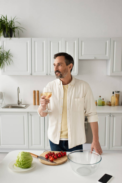 uomo barbuto in camicia bianca in possesso di un bicchiere di vino e guardando lontano vicino a pomodori ciliegini maturi e lattuga fresca - Foto, immagini