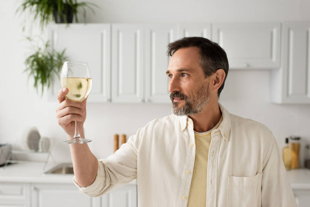 smiling man with grey beard looking at glass of white wine in kitchen - Foto, afbeelding