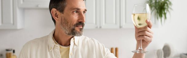 bearded man in white shirt looking at glass of white wine in kitchen, banner - Foto, Bild