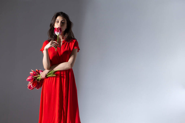 Переднее изображение привлекательной брюнетки в красном платье, держащей букет красных тюльпанов и запахов, изолированный серый фон. Пространство для текста. - Фото, изображение