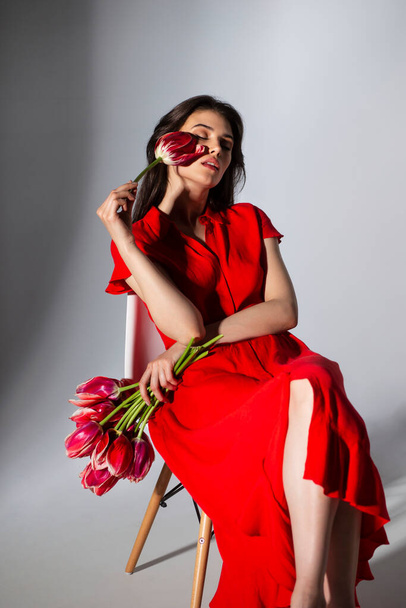 Elegáns barna szexi fiatal nő piros ruhában kezében egy gyönyörű piros tulipán, ül a széken, elszigetelt szürke háttér. - Fotó, kép