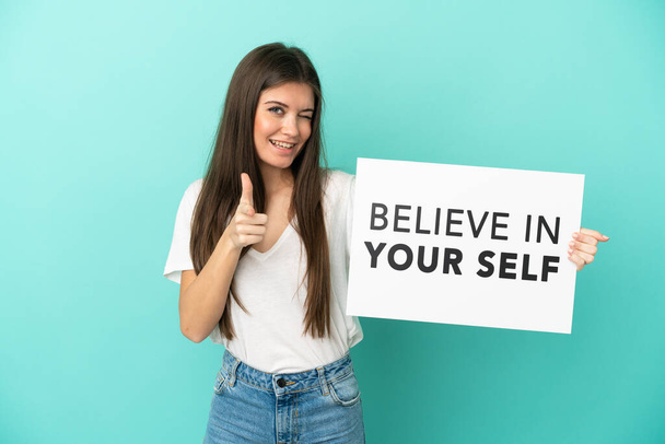 Mladá běloška izolovaná na modrém pozadí drží plakát s textem Věřte ve své já a ukazujte dopředu - Fotografie, Obrázek