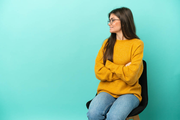 Young caucasian woman sitting on a chair isolated on blue background with arms crossed and happy - Photo, Image