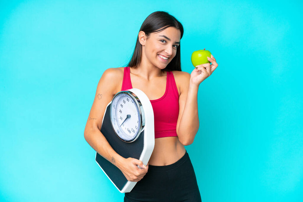 Young caucasian woman isolated on blue background with weighing machine and with an apple - Foto, Bild
