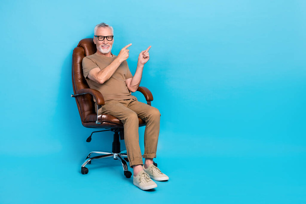 Full size photo of cheerful person sit chair indicate fingers empty space isolated on blue color background. - Foto, immagini
