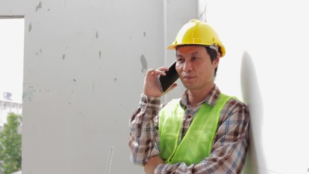 Строитель в жёлтом шлеме держит смартфон, разговаривающий с координатором на стройке. Концепция строительной отрасли. проект недвижимости - Кадры, видео