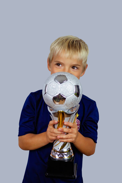 Träume werden wahr. Junger Fußballer im blauen Trikot küsst nach dem Siegtor im Fußballturnier einen Siegerpokal, isoliert vor hellem Hintergrund. Sport, Sieger und Erfolg. - Foto, Bild