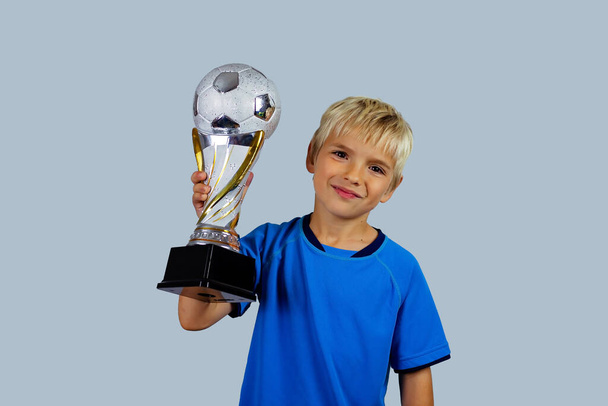 Mavi formalı genç futbolcu, hafif arka planda izole bir şekilde futbol maçının galibiyet golünü attıktan sonra kupa kaldırıyor. Spor, kazanan ve başarı. Hayaller gerçek olur. - Fotoğraf, Görsel