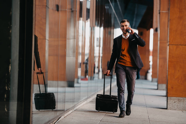 Going to airport terminal. Confident businessman traveler walking on city streets and pulling his suitcase drinking coffee and speaking on smartphone. High quality photo - Фото, зображення