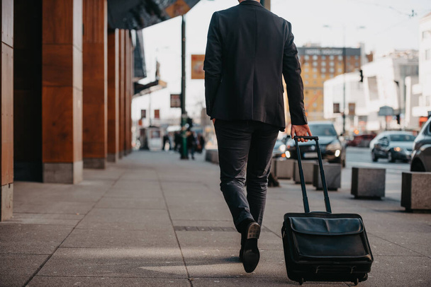 Going to airport terminal. Confident businessman traveler walking on city streets and pulling his suitcase drinking coffee and speaking on smartphone. High quality photo - Foto, Bild
