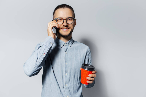 Studioporträt eines jungen lächelnden Mannes, der auf einem Smartphone spricht und in einer anderen Hand, auf weißem Hintergrund, eine rote Papptasse zum Mitnehmen hält. Mit blauem Hemd und Brille. - Foto, Bild