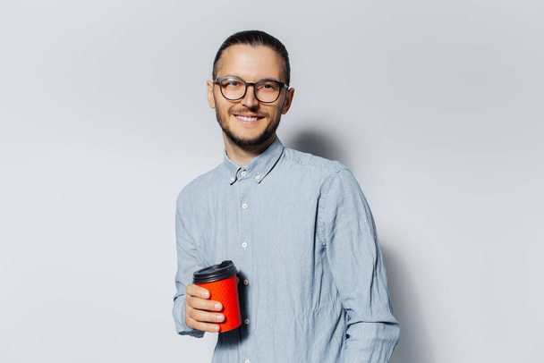Studioporträt eines jungen lächelnden Mannes mit roter Papptasse zum Mitnehmen in der Hand, auf weißem Hintergrund. Mit blauem Hemd und Brille. - Foto, Bild