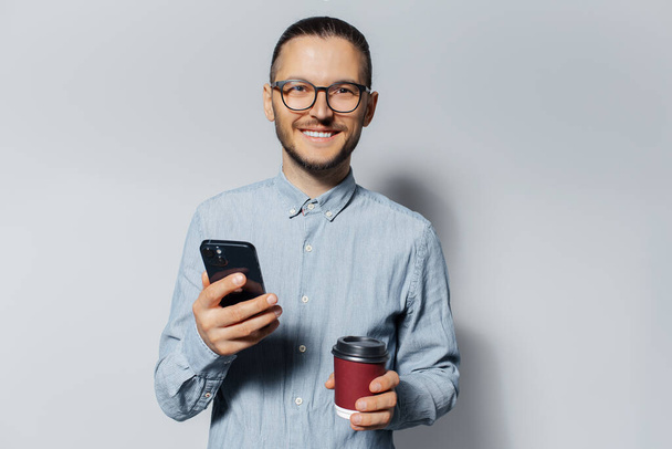 Studio portrait of young smiling man holding a smartphone and red paper cup for coffee takeaway in hands on white background, wearing blue shirt and eyeglasses. - Photo, Image