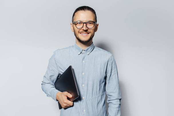 Studio portrait of young smiling man with laptop in hand, on white background. Wearing blue shirt and eyeglasses. - 写真・画像