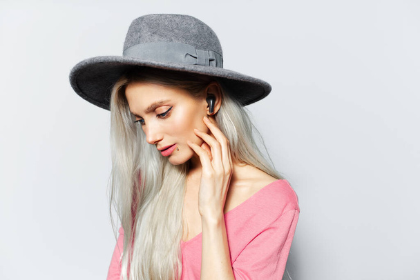 Studio portrait of young blonde cute girl listening the music via earbuds, on white background, wearing grey hat and pink shirt. - Foto, Imagen