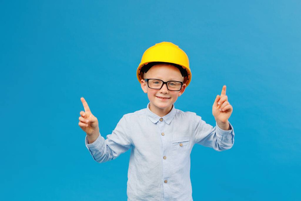 Милый мальчик-строитель в жёлтой каске, стоящий в крытой студии на синем фоне и указывающий пальцами на направление. Будущий строитель - Фото, изображение