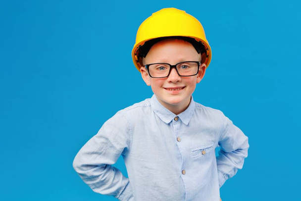 Cute boy construction worker in yellow hard hat standing in indoor studio on blue background. Future construction worker - Foto, Imagem
