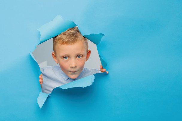 Happy cute boy is having fun played on blue background wall, climbs through a hole in the paper. Bright and funny emotions of the boy - Photo, image