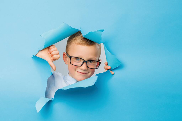 Happy cute businessboy in eyeglasses is having fun on blue background wall, climbs through a hole in the paper. Bright and funny emotions of the boy - Valokuva, kuva