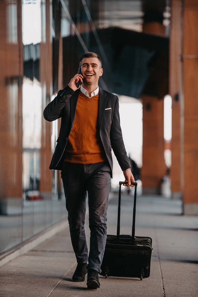 Going to airport terminal. Confident businessman traveler walking on city streets and pulling his suitcase drinking coffee and speaking on smartphone. High quality photo - Zdjęcie, obraz