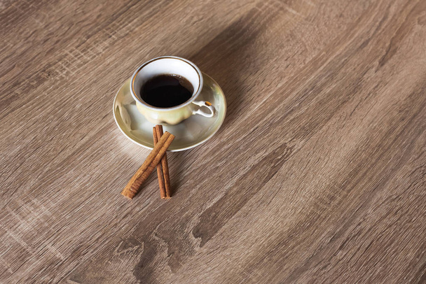 A cup of hot aromatic coffee in a saucer, yellow, stands on a wooden textured brown table, decorated with cinnamon sticks. - Foto, Bild