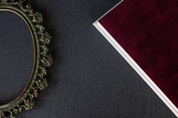 A velvet burgundy photo book endpaper with white pages and a copper vintage baroque frame lie on a dark gray fabric indoors. - Photo, Image