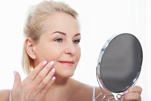 Happy middle aged woman model touching face skin looking in mirror. Smiling mature older lady enjoying healthy skin care. Aging beauty, skincare treatment cosmetic products concept. - 写真・画像