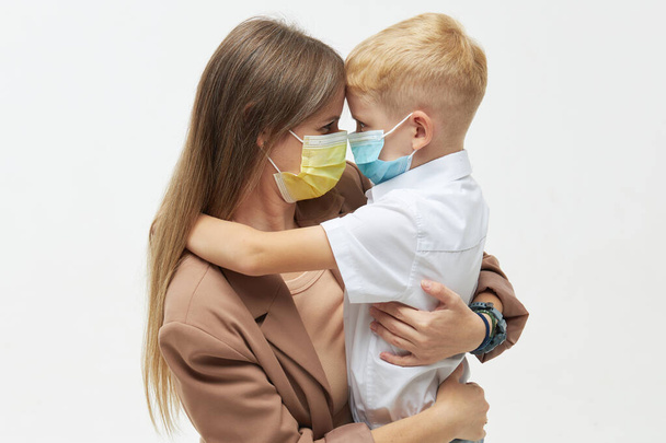 Caring for loved ones. Young mother hugs little son in protective medical masks during Covid-19 pandemics. Studio white background - Фото, изображение