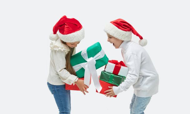 Christmas child. Cute toddler girl abd boy in santa claus caps holding high stack of colorful gift boxes on white. Preparing for New Year's Eve in Italy - Photo, Image