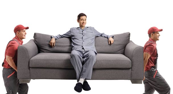 Movers carrying a sofa with a man in pajamas sitting on the sofa isolated on white background - Photo, Image