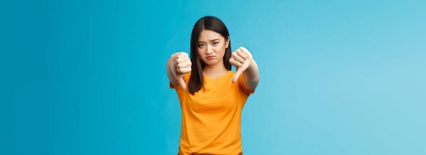 Upset gloomy asian girl with dark haircut frowning sulking sad, disappointed, show thumbs-down grimacing, give negative opinion, judging bad uninteresting movie, stand blue background. - 写真・画像