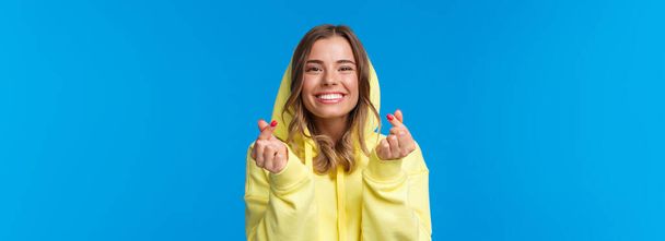 Retrato de cerca de la linda y tonta chica rubia caucásica con capucha amarilla, mostrando el gesto del corazón coreano y sonriendo, vibrando, disfrutando de la fiesta, divirtiéndose sobre el fondo azul. Copiar espacio
 - Foto, imagen