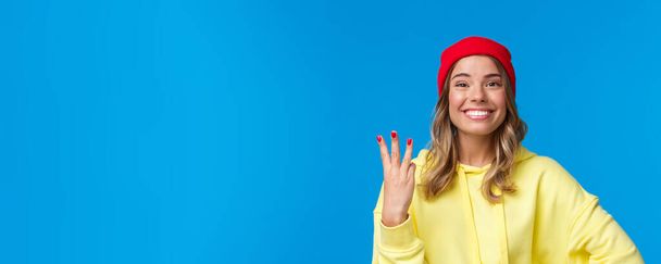Mujer caucásica de buen aspecto en gorro rojo y sudadera con capucha amarilla, mostrando el número tres y sonriendo, hacer orden o reserva, de pie orgulloso y encantado, fondo azul. Copiar espacio
 - Foto, Imagen