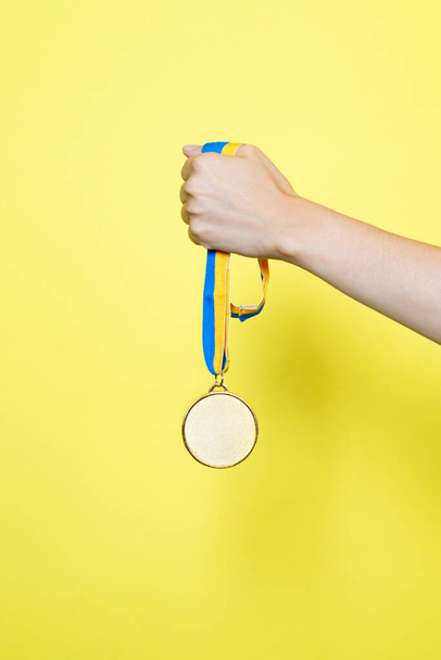 Hand hold Gold medal on yellow blue ribbon isolated on yellow background. Vertical Medal mockup. - Foto, Imagen