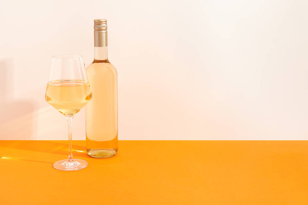 Μπουκάλι και ποτήρι λευκό κρασί σε begie και πορτοκαλί φόντο με βαθιές σκιές. Mock up ποτό με θέση για σας lable και κείμενο. Σχεδιασμός εμπορικού σήματος συσκευασίας προϊόντος. - Φωτογραφία, εικόνα