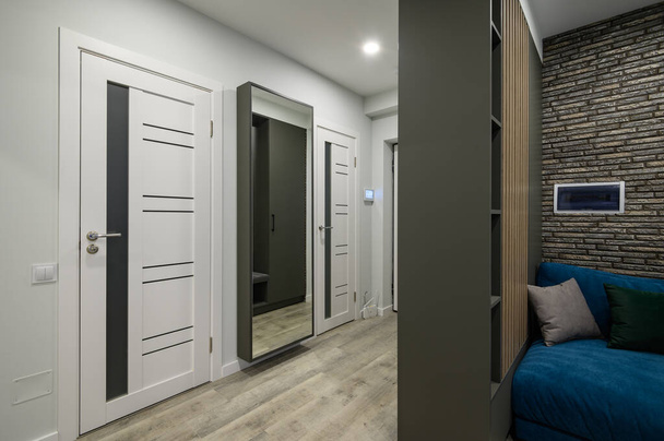 Entree hal bevestigd aan kleine moderne woonkamer met grote blauwe comfortabele bank achter de kledingkast, deur naar badkamer en spiegel op de achtergrond - Foto, afbeelding
