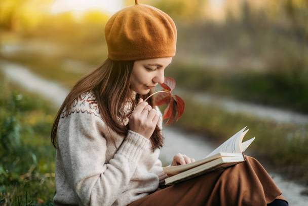 Πορτραίτο μιας γυναίκας σε ένα γαλλικό μπερέ. Μια νεαρή κοπέλα με πλεκτό πουλόβερ και καφέ μπερέ κάθεται στο γρασίδι διαβάζοντας ένα βιβλίο. Γαλλίδα. Χιλιετής γενιά. - Φωτογραφία, εικόνα