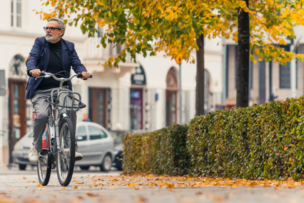 Outdoor shot. Cycling concept. Full-length shot of smart-dressed caucasian man in his 60s with gray hair riding city bike in park where leaves are changing their color to yellow. High quality photo - Zdjęcie, obraz