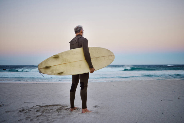 Surfer, surfboard and senior man on beach at sea waves in during sunset during summer vacation in Hawaii. Professional male athlete rest after training or practice surfing sport outdoor at the ocean. - Foto, Imagem