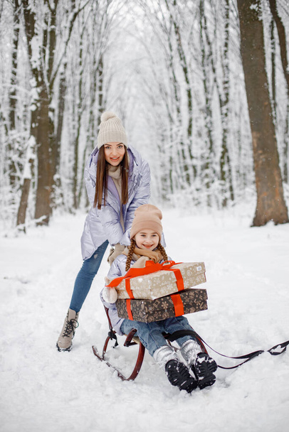 Mother with her child having fun in forest on snow day. Girls wearing purple warm jackets and knitted hats. Woman driving her daughter on a sled. - Photo, image