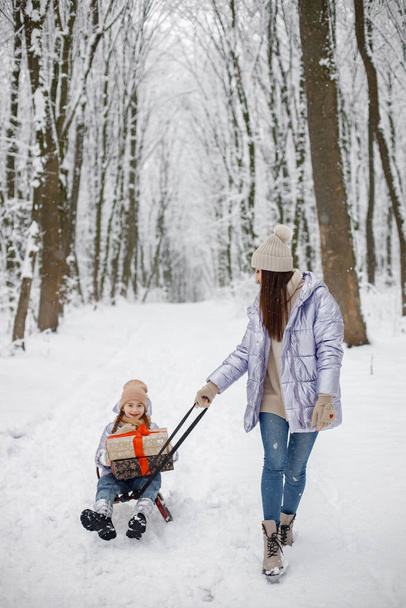 Mother with her child having fun in forest on snow day. Girls wearing purple warm jackets and knitted hats. Woman driving her daughter on a sled. - 写真・画像