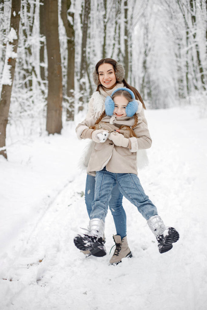 Mother playing with her child and holding her on hands in forest on a winter day. Girls wearing beige warm clothes, jeans and earmuffs. Woman and her daughter posing for a photo. - Foto, Imagem