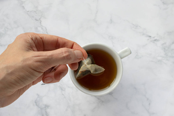Person holding a tea bag. Teabag in woman hand. Cup of green herbal tea on white marble table background. Preparing afternoon tea process. Teatime. Top view - Photo, Image