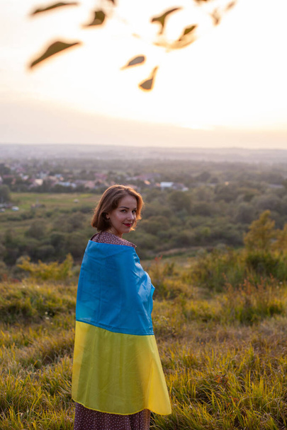 長いドレスで幸せな女性は、日没の背景にウクライナの黄色と青の旗を自分自身をカバーしています。戦争に対するウクライナ人。独立記念日。旗の日。憲法の日だ。後景. - 写真・画像