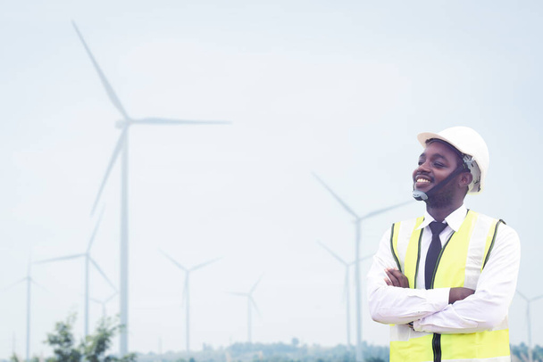 Αφρικανός μηχανικός άνθρωπος στέκεται με χαμόγελο μπροστά από τις ανεμογεννήτριες παραγωγής ηλεκτρικής ενέργειας.Έννοια της ανάπτυξης βιωσιμότητας από εναλλακτικές πηγές ενέργειας - Φωτογραφία, εικόνα