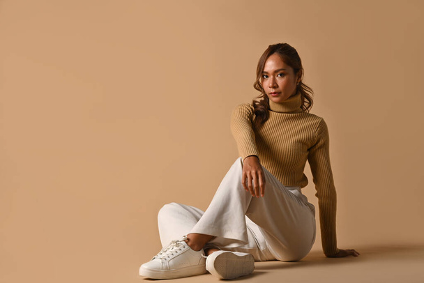 流行のセーターを茶色の背景に座って若い女性の完全な長さ。ファッションスタジオの写真、秋と冬のコンセプト. - 写真・画像