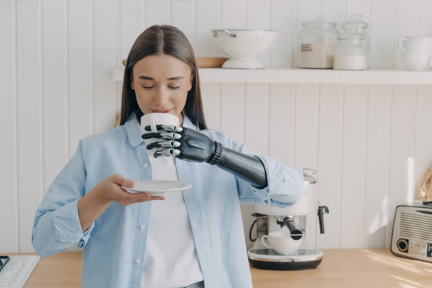 Девушка-инвалид пьет кофе или чай, держа кружку высокотехнологичным бионическим протезом руки в уютной кухне. Молодая женщина с ограниченными возможностями наслаждается утренней рутиной дома с помощью искусственной руки. - Фото, изображение