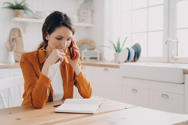 Puzzled woman making phone call, taking notes sitting at kitchen table. Pensive female freelance worker consulting client or discussing project with colleague at home. Remote job concept. - Фото, зображення